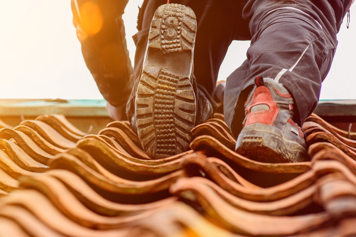 Travailleur en pleine pose de tuiles sur un toit, vu de près avec ses chaussures de sécurité sur les tuiles ondulées, sous une lumière de fin de journée.